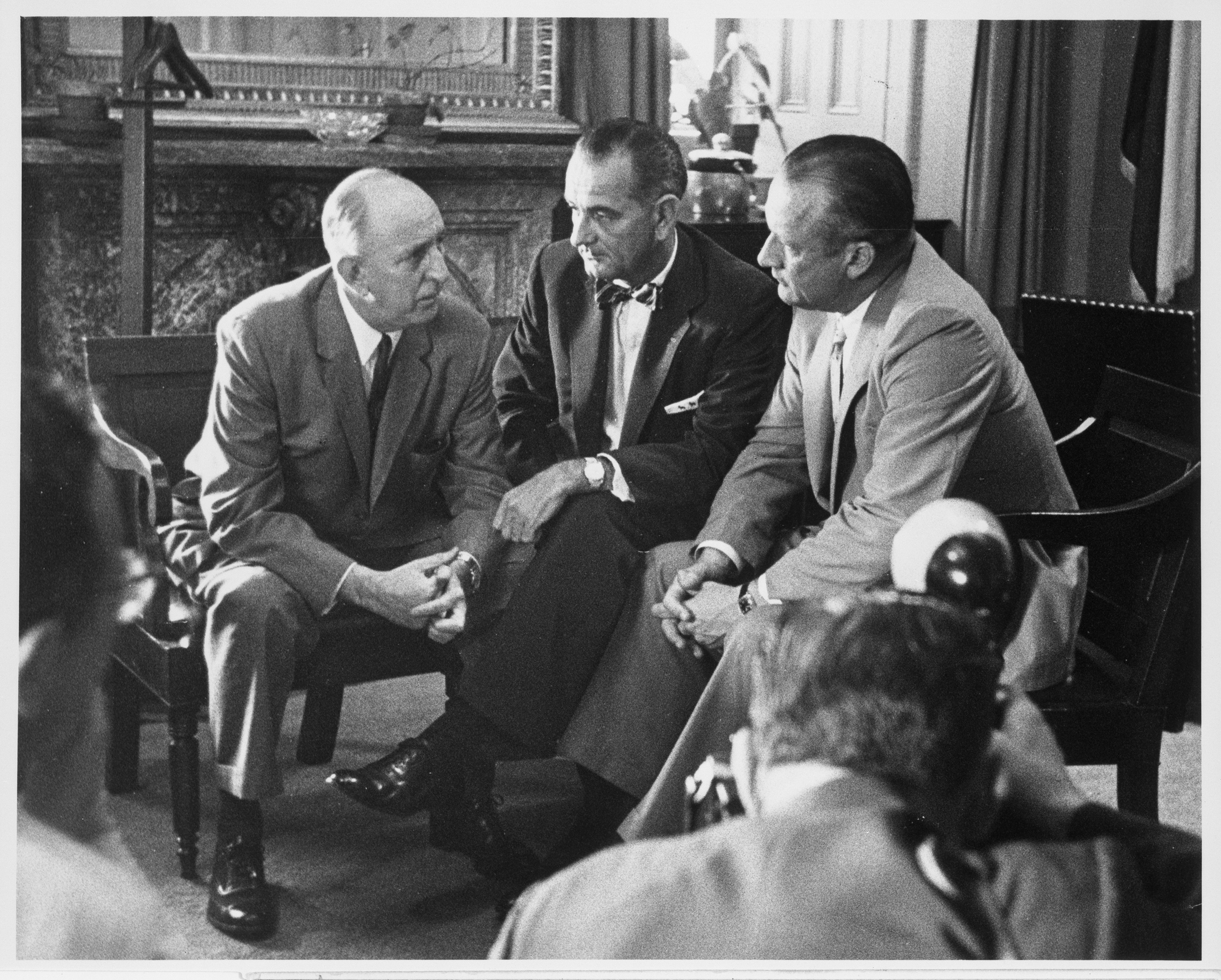 Three men in suits sit in deep conversation with others looking on. 